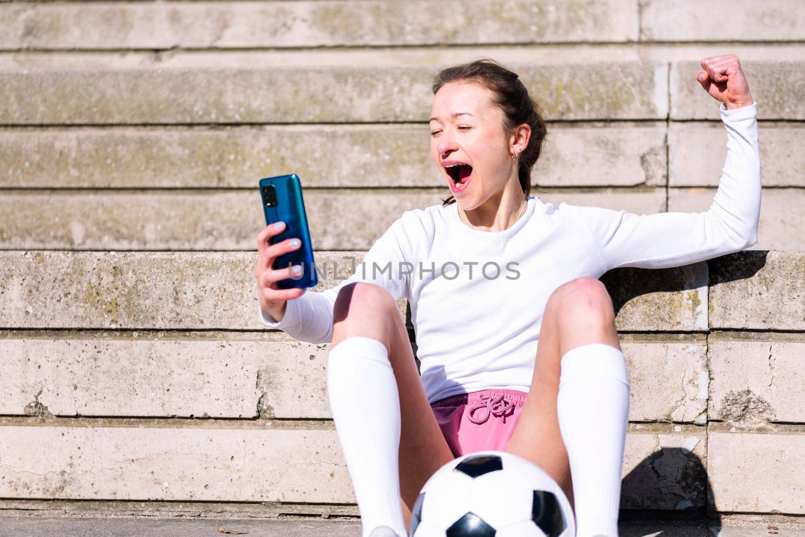 female football fan celebrates a victory when she is watching her mobile phone, concept of technology and urban soccer lifestyle, copy space for text