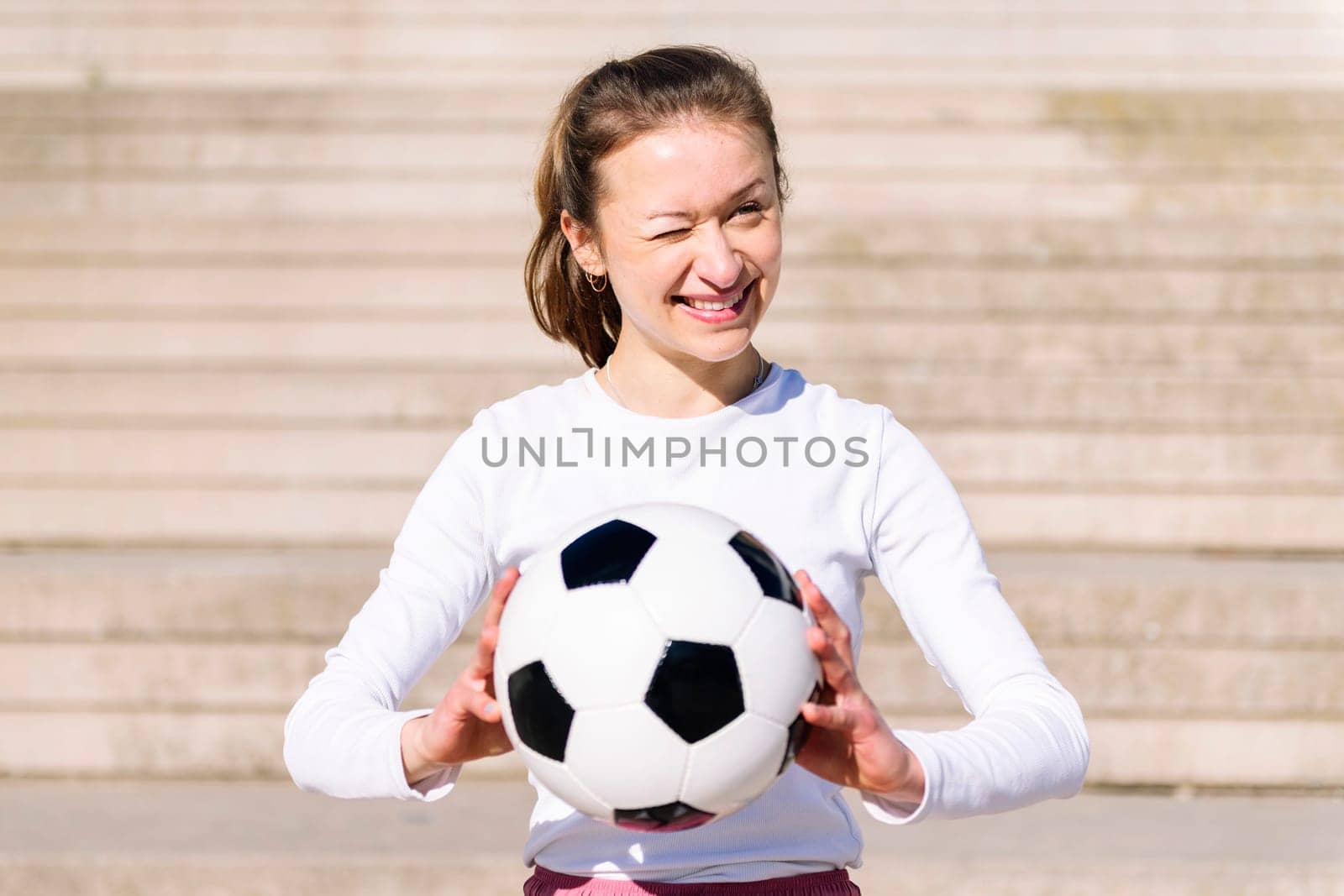 portrait of a smiling sports woman winking in a concrete soccer court with a ball in her hands, concept of football lifestyle and urban sport in the city