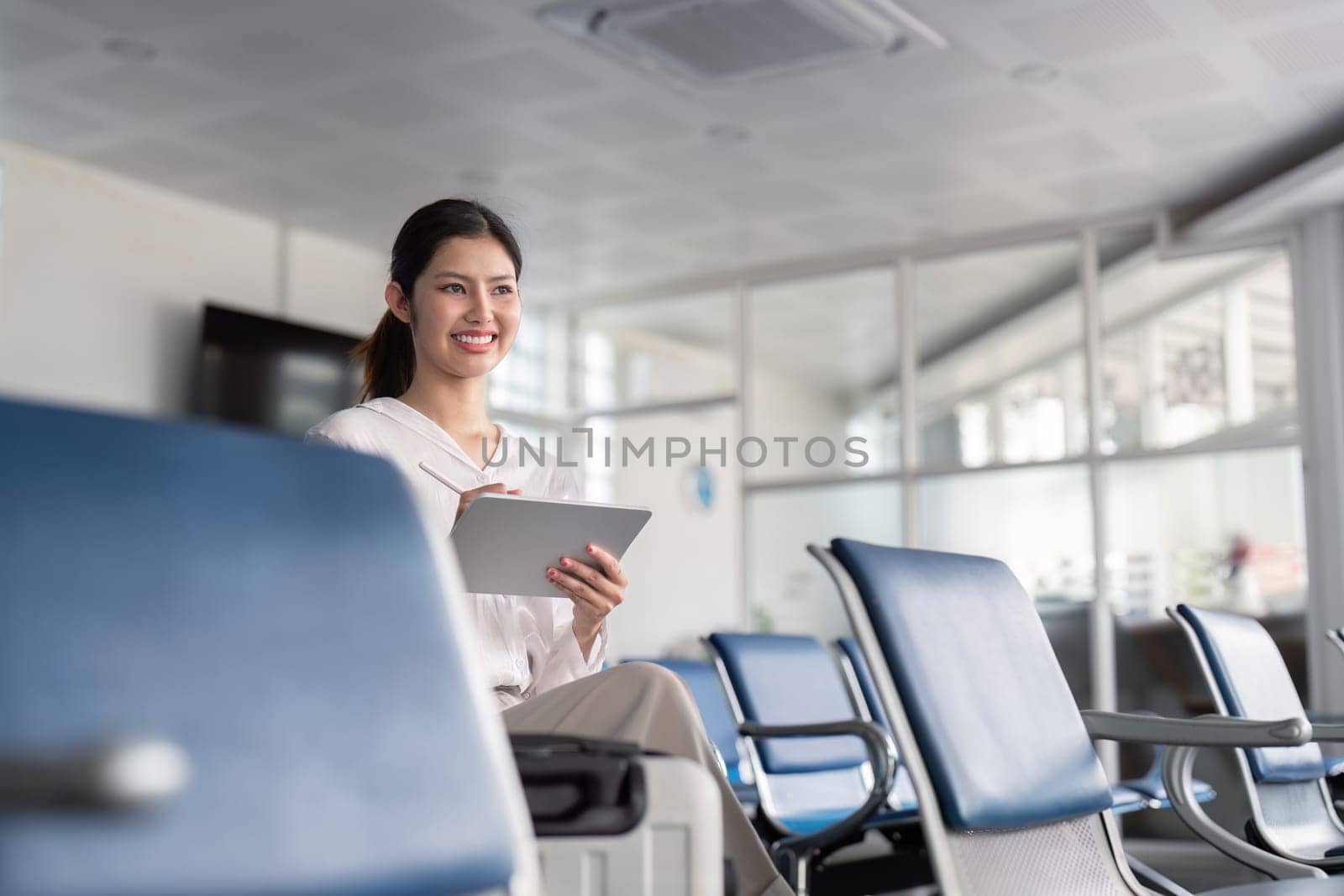 A business professional using a tablet in a modern airport lounge, featuring glass partitions and comfortable seating, ideal for business travel and productivity.