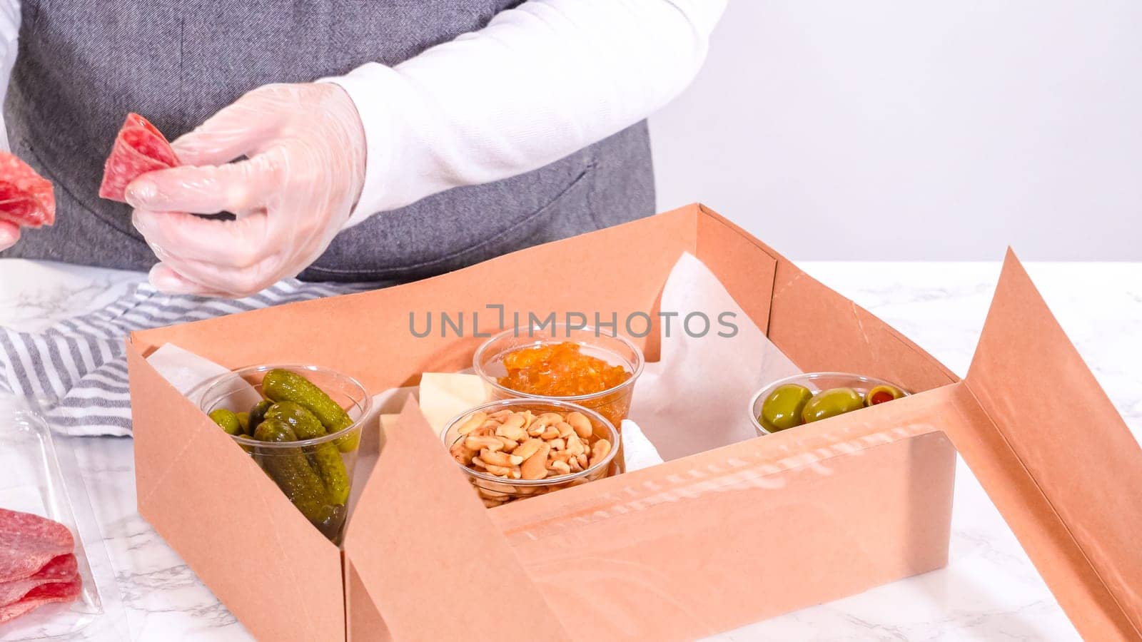 Hands are meticulously adding fresh red grapes to a bowl, complementing a beautifully arranged charcuterie box brimming with a variety of cheeses, olives, and cured meats, set against a sleek marble surface.