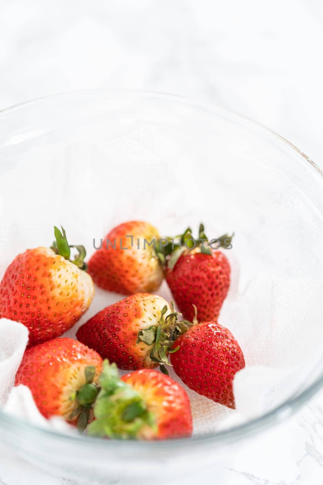 Bright red strawberries, interspersed with signs of mold, rest in a glass bowl lined with a paper towel on a white napkin, indicating improper storage techniques.