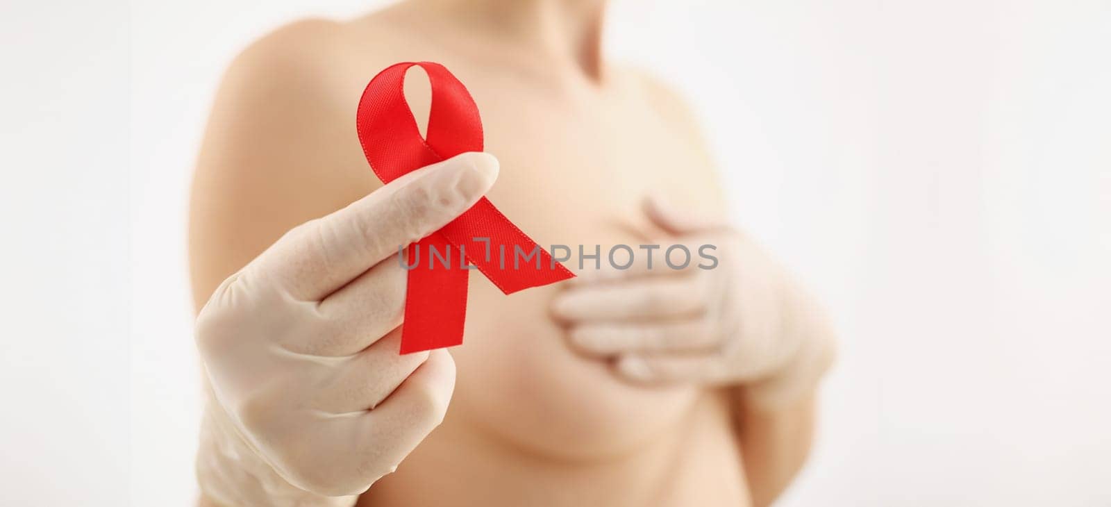 Young woman holding red ribbon in her hand and covering her breasts closeup. Prevention of breast cancer concept
