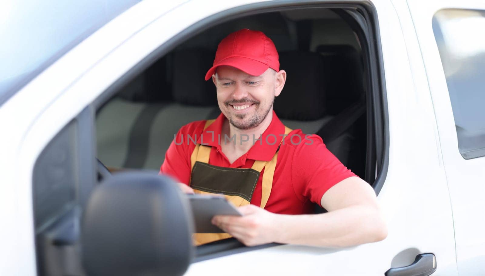 Man courier in uniform sitting at wheel of car with digital tablet in his hands by kuprevich