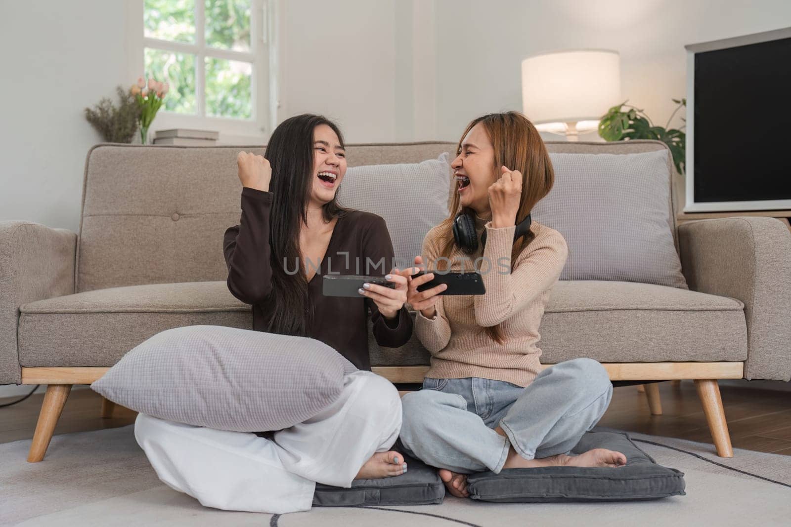 Happy lesbian couple having fun playing mobile games together in a cozy living room, celebrating and enjoying their time.