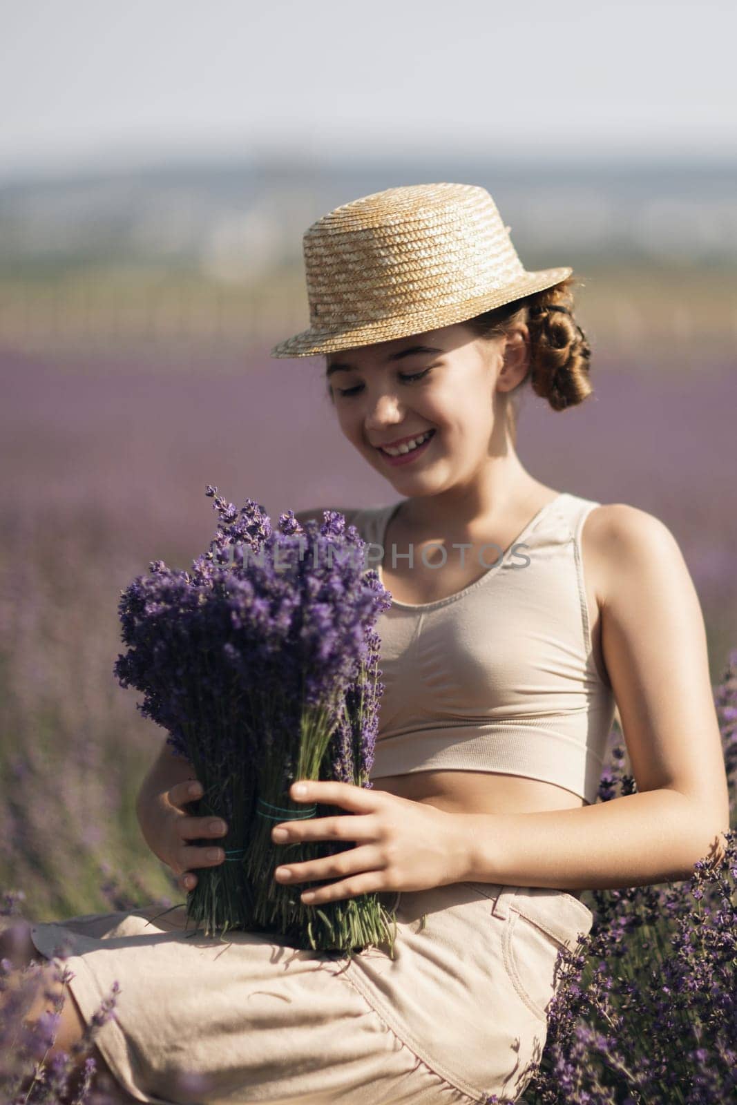 A woman is sitting in a field of lavender flowers and wearing a straw hat. She is smiling and holding a bouquet of flowers. Scene is peaceful and serene, as the woman is surrounded by the beauty of nature.