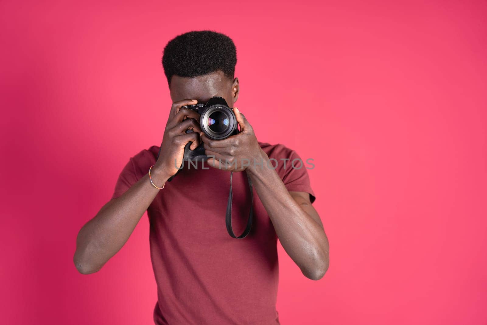 Young black man taking a photograph with a mirrorless camera, wearing a red t-shirt on a carmine red background with copy space. by Ceballos