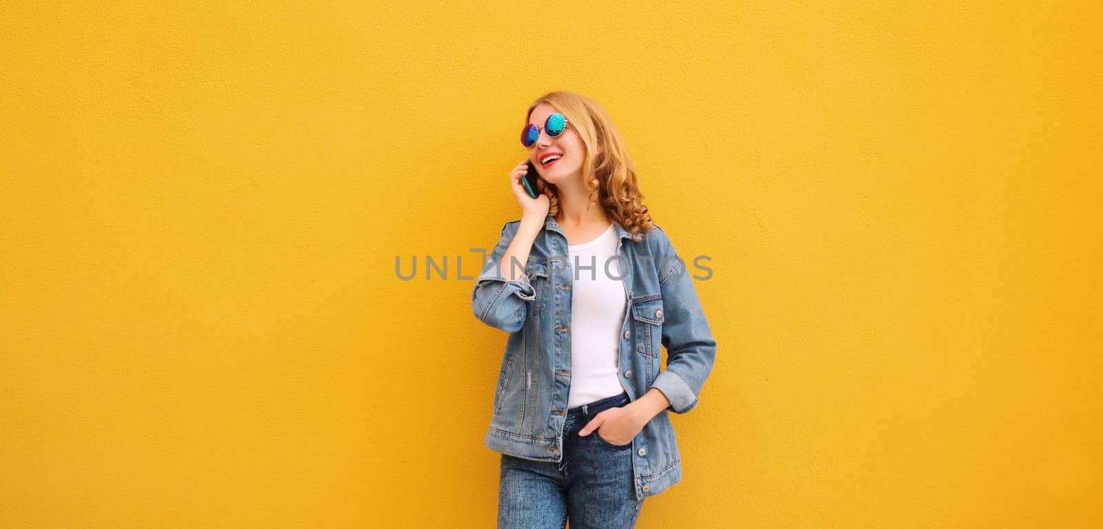 Happy cheerful young woman laughs calling on phone, girl talking on the smartphone in denim clothing looks away on bright yellow background