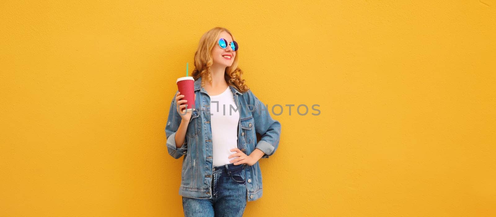 Summer vacation, happy joyful young woman with cup of coffee, stylish modern girl in denim jacket drinking fresh juice looking away posing on vivid yellow wall background