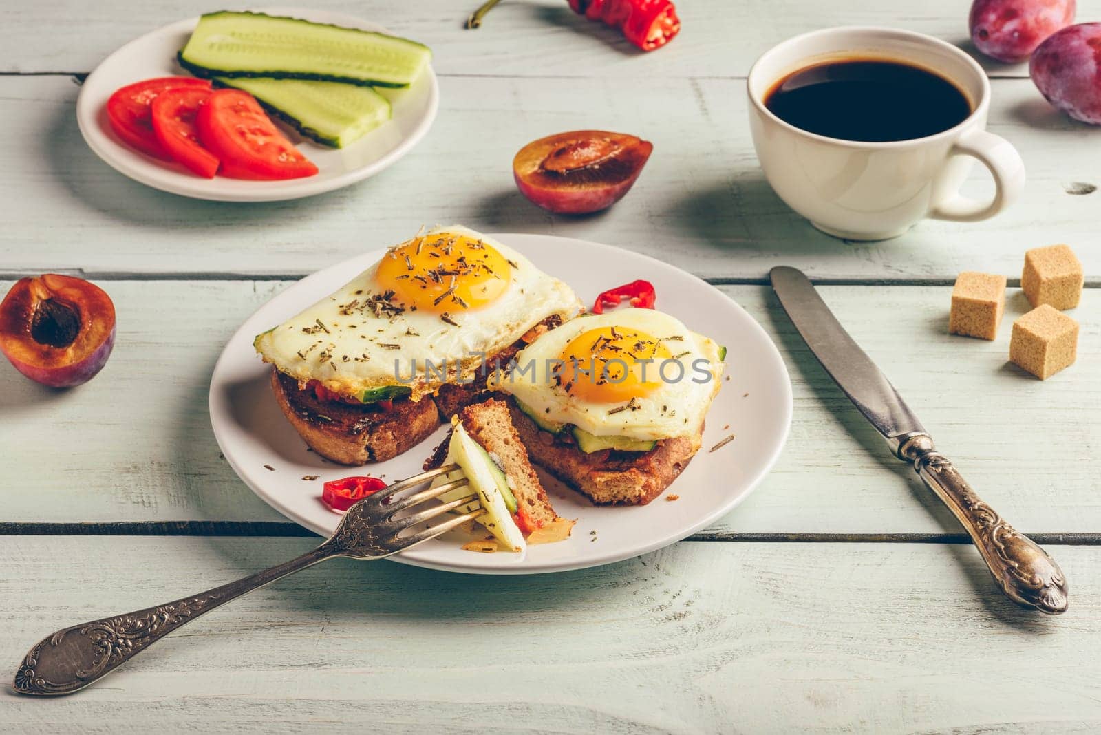 Sandwiches with vegetables and fried egg on white plate, cup of coffee and some fruits over wooden background.