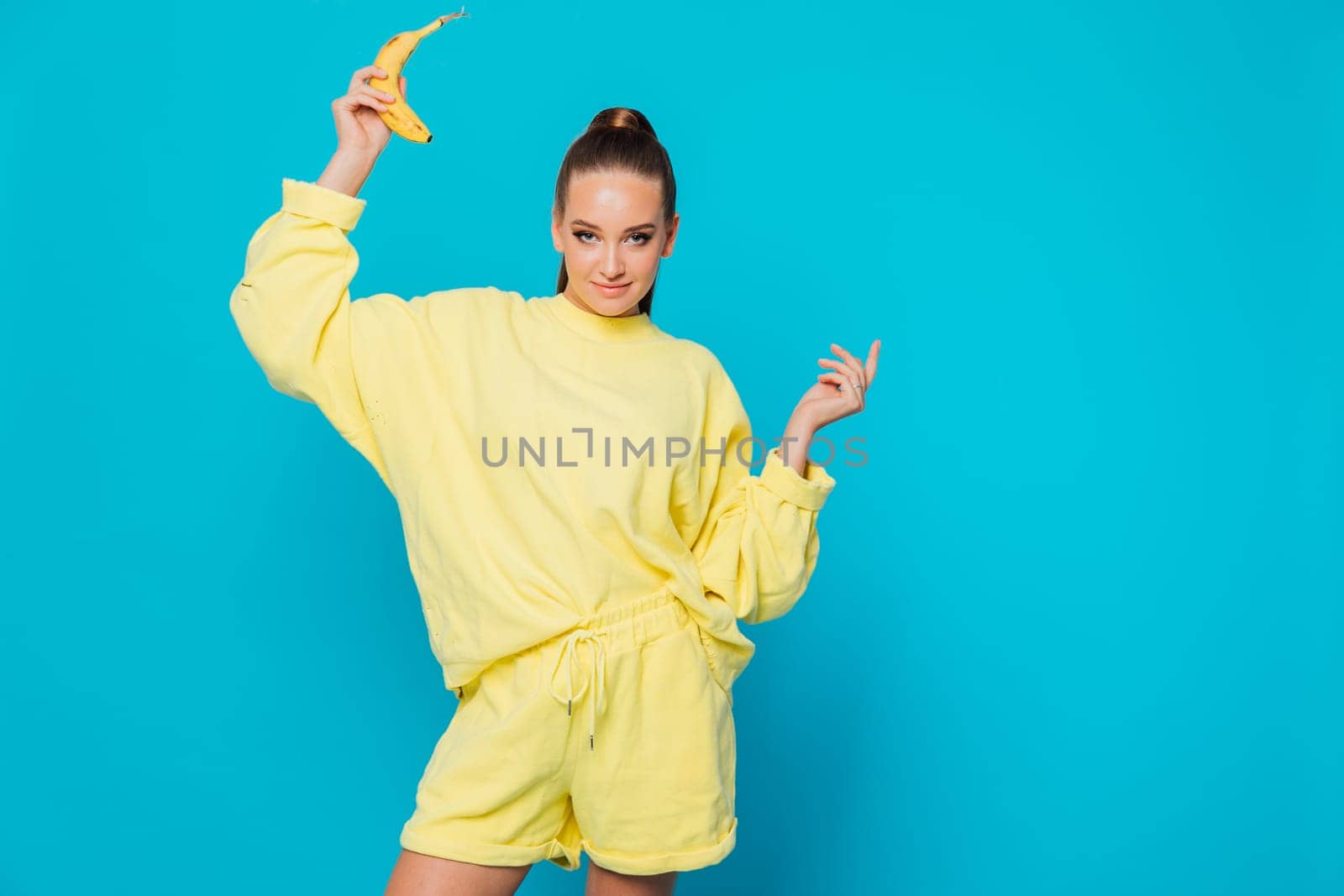 a woman in a yellow suit stands on a blue background with a banana in her hand