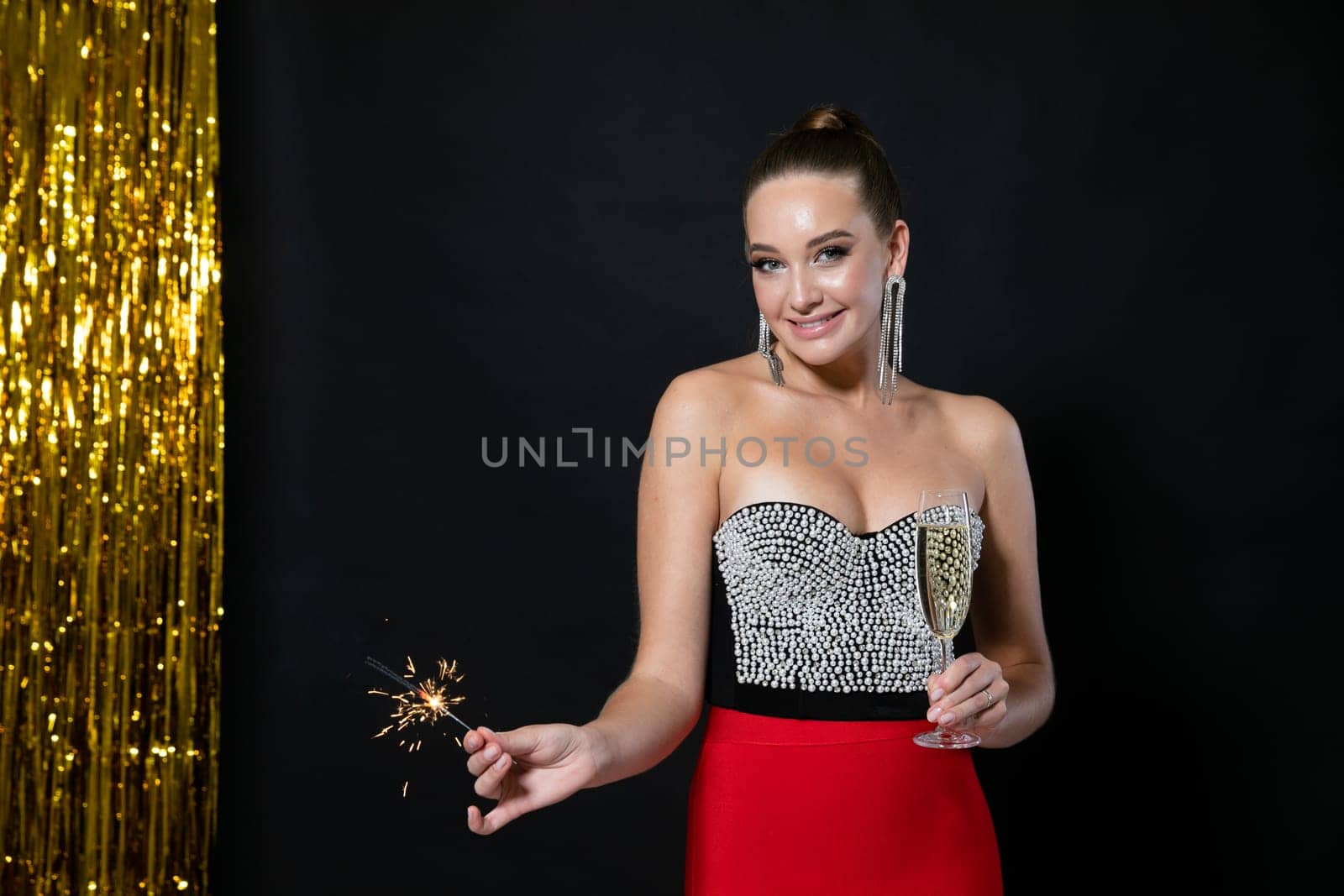 a beautiful woman in a festive dress stands with a glass of champagne and a sparkler at the black background with tinsel
