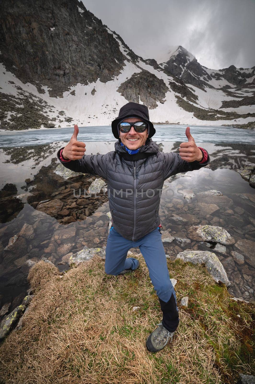 A young joyful man in black sunglasses and sportswear stands in front of a mountain lake and a mountain range. Happy tourist looking at camera, smiling and thumbs up showing class gesture.