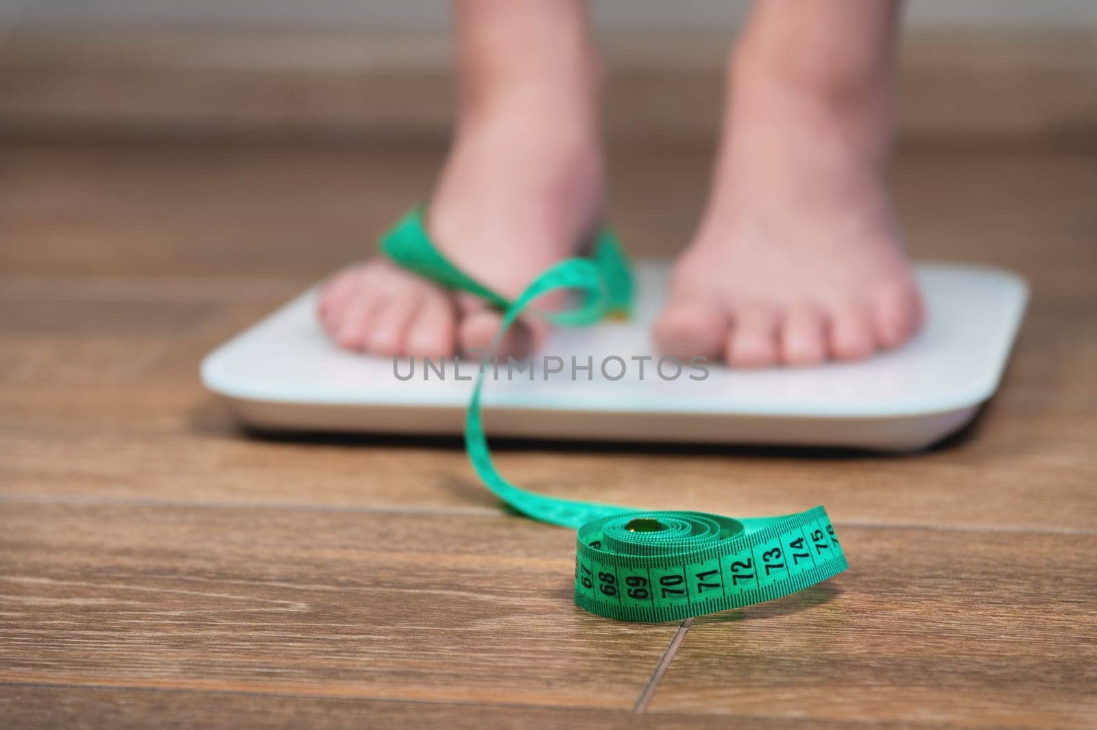 A girl with swollen feet uses a scale next to a measuring tape on a wooden floor. Unrecognizable woman weighing herself.