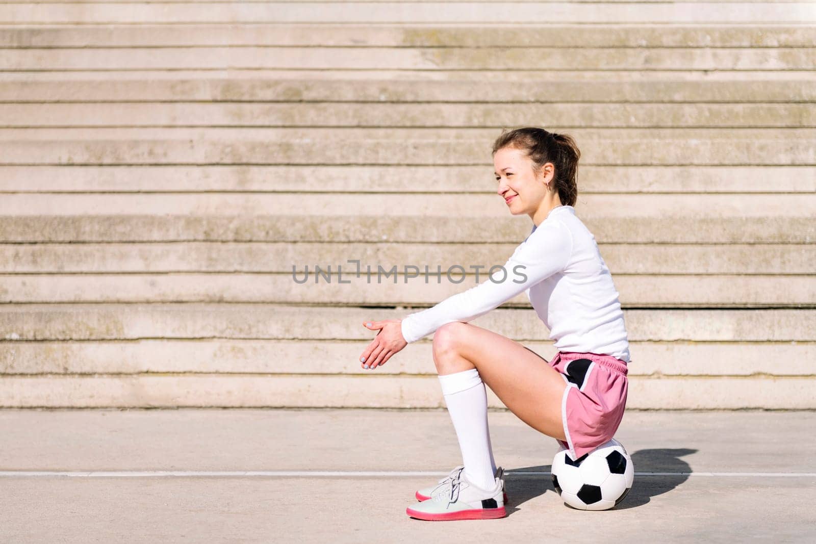smiling sports woman in a concrete soccer court sitting on her soccer ball, concept of football lifestyle and urban sport in the city, copy space for text