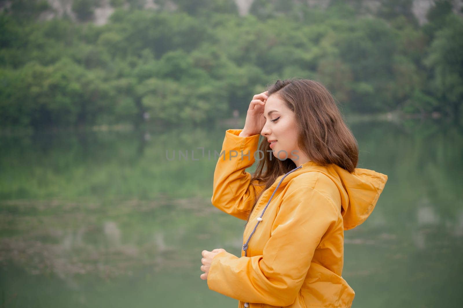 A woman in a yellow jacket is standing by a lake. She is looking at the water and she is lost in thought