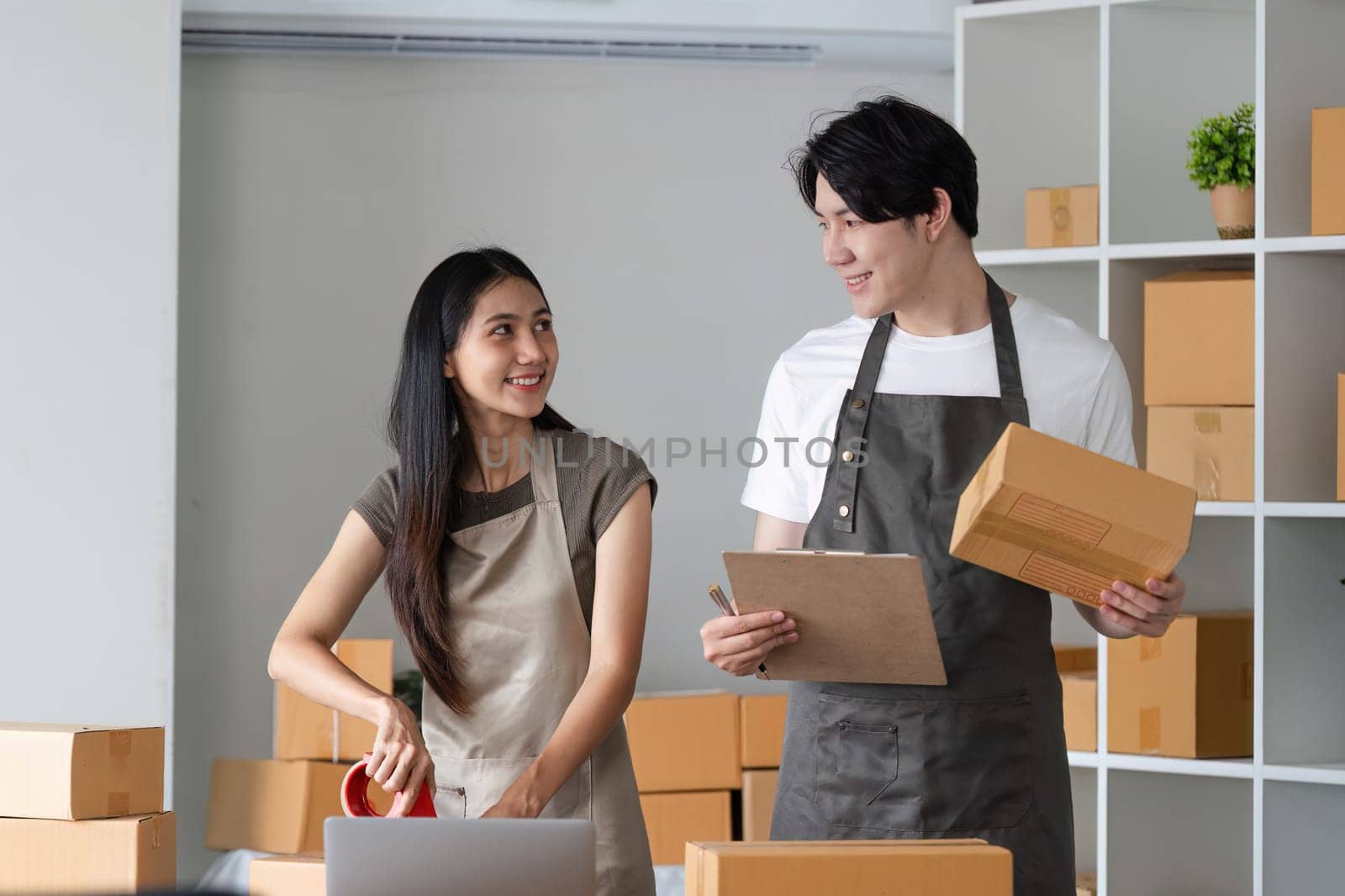 A young couple in aprons taking online orders and packing products in a modern home office, surrounded by cardboard boxes and a laptop.