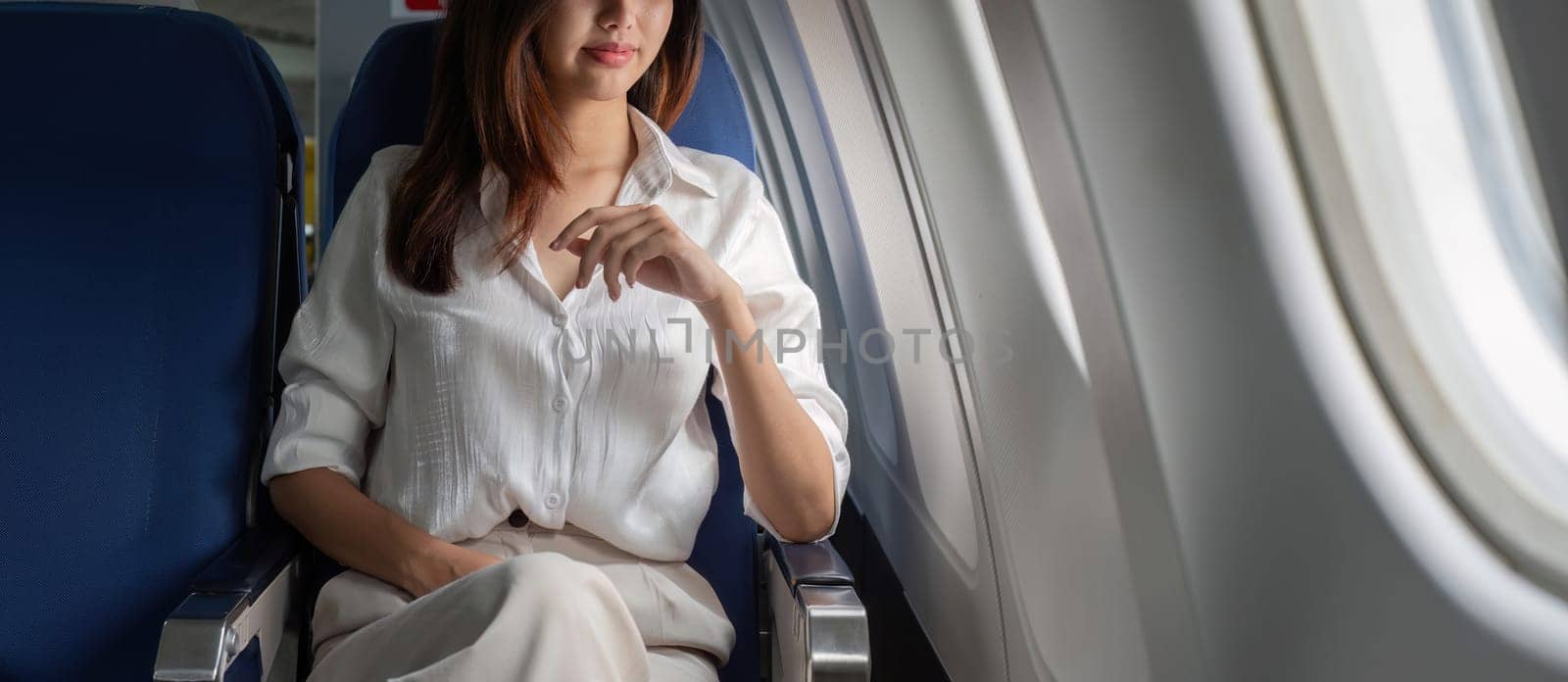 Young businesswoman seated in airplane cabin, wearing a white blouse, looking out the window, representing modern professional travel lifestyle.