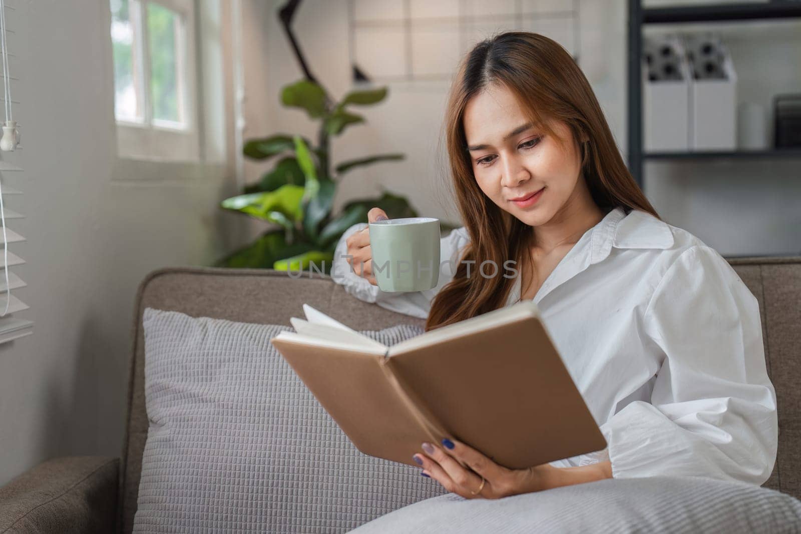 A young woman starts her day with a relaxing morning routine, enjoying a cup of coffee and reading a book in a cozy, modern living room.