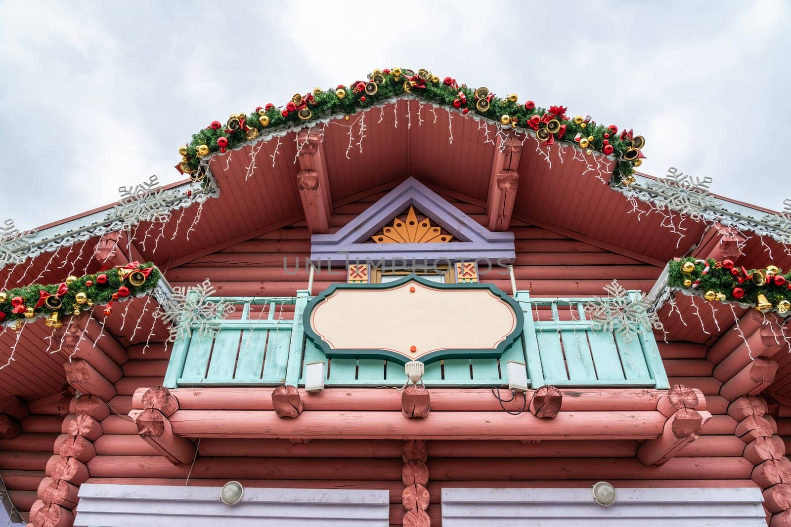 This photo shows the exterior of a log cabin building adorned with Christmas decorations, including garlands of evergreen branches, red and gold ornaments, and snowflakes.