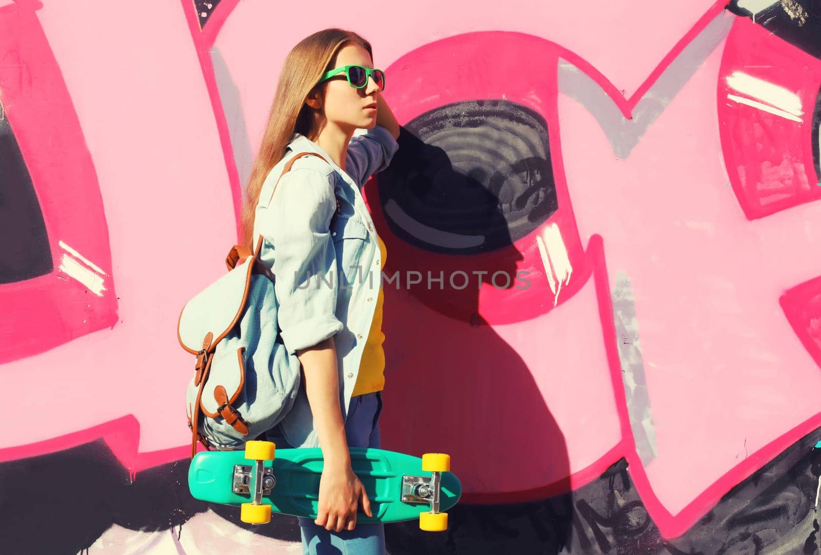 Stylish young girl with skateboard in glasses, modern teenager girl looking away posing against the background of pink graffiti wall