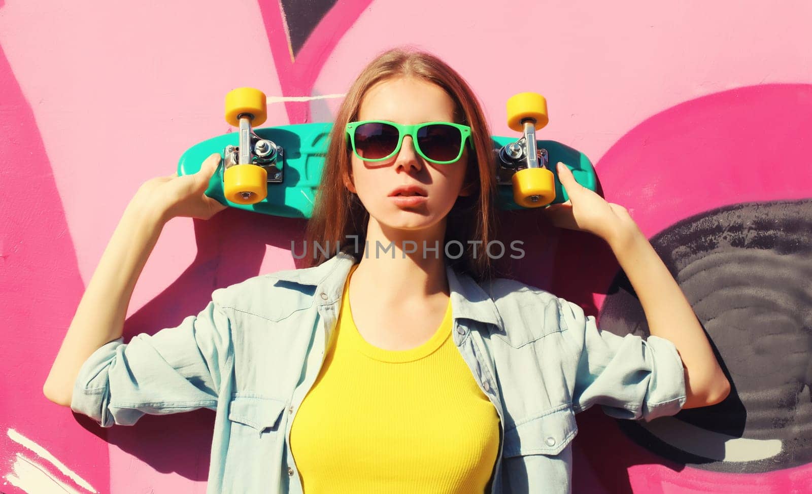 Portrait of stylish young girl with skateboard in glasses, modern teenager girl posing against the background of pink graffiti wall
