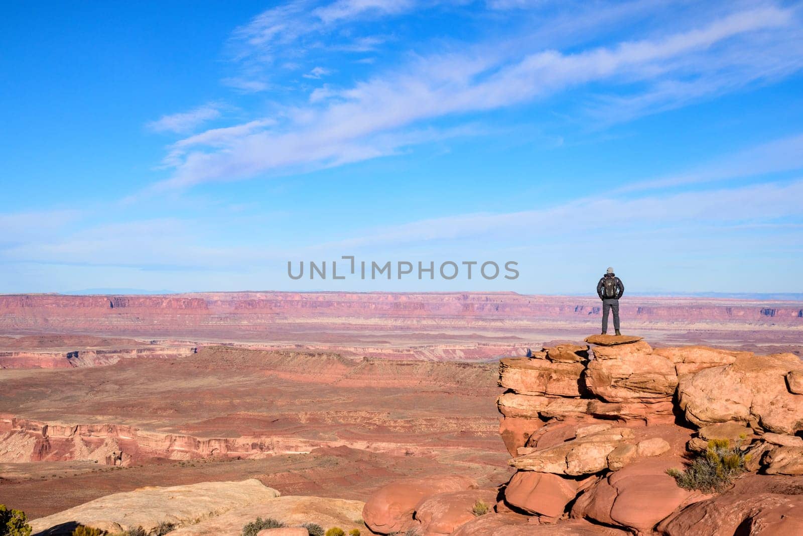 A solitary man stands on a rock overlook, taking in the expansive views of the dramatic Canyonlands in Utah. The clear blue sky enhances the vibrant red rock formations, symbolizing adventure - USA