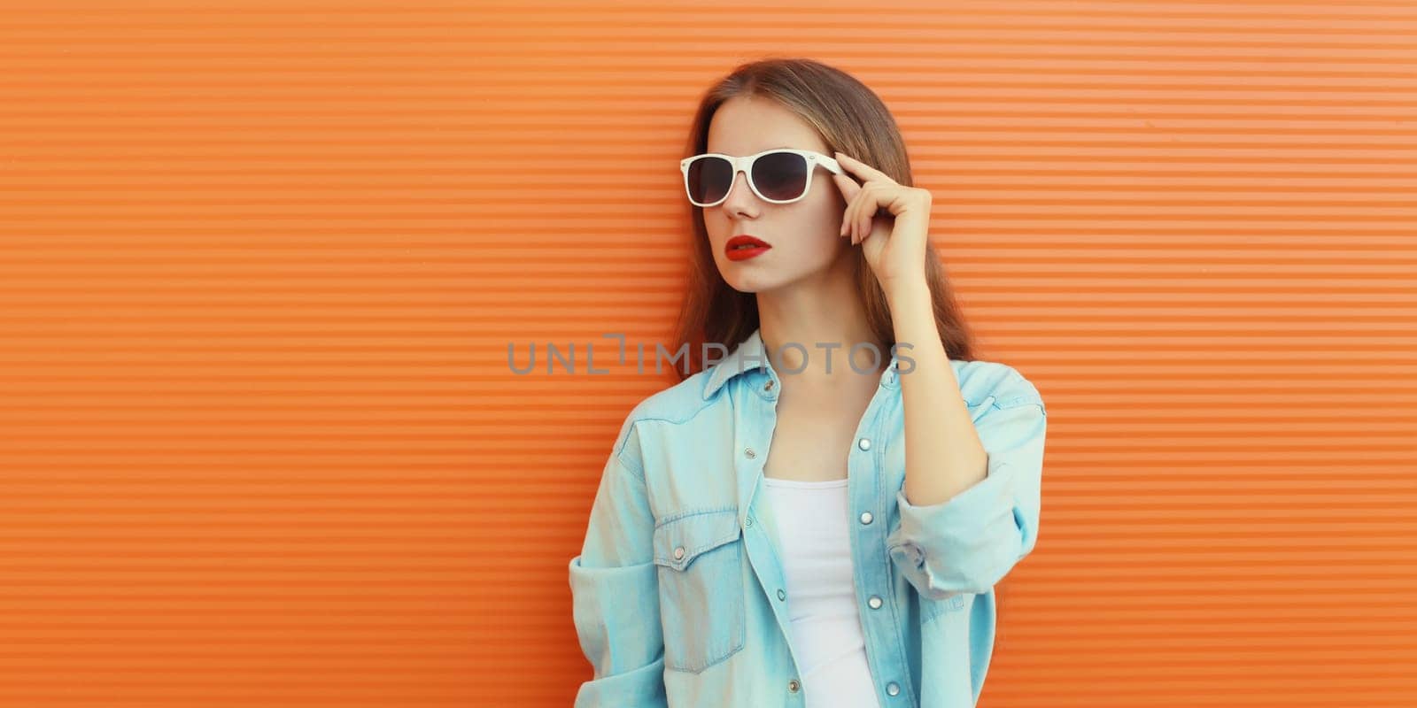 Portrait of modern stylish young woman in white glasses, casual clothing posing on vivid orange wall background