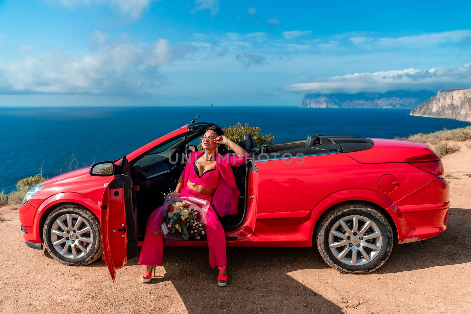 A woman is sitting in the back of a red convertible car. The car is parked on a dirt road near the ocean. The woman is wearing a pink suit and red shoes. Scene is relaxed and carefree