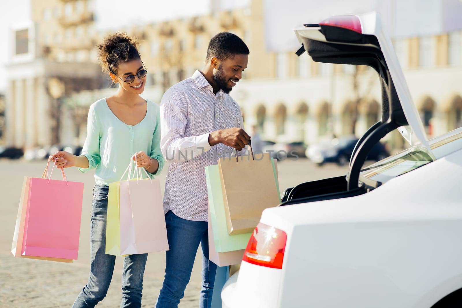 Black couple is loading shopping bags into the trunk of their car after a day of shopping. The woman is holding a pink shopping bag in her left hand and a multi-colored bag in her right.