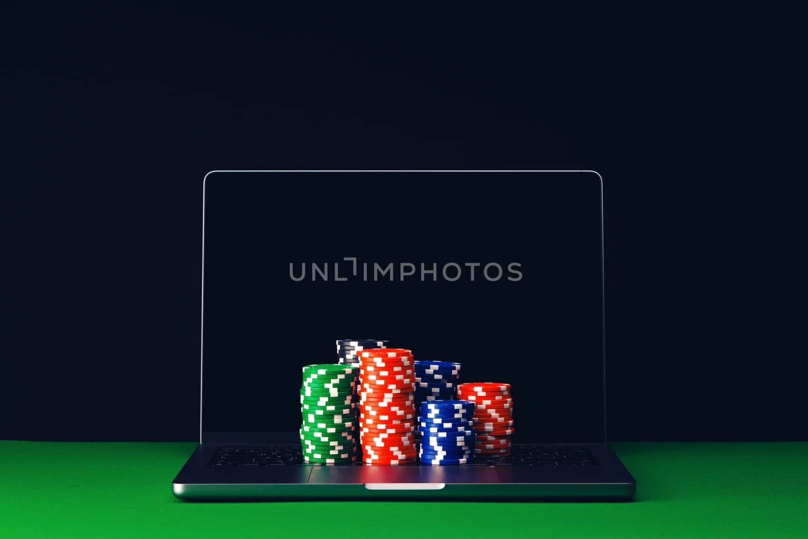 A laptop computer rests on a green felt surface with several stacks of poker chips in front of the screen.