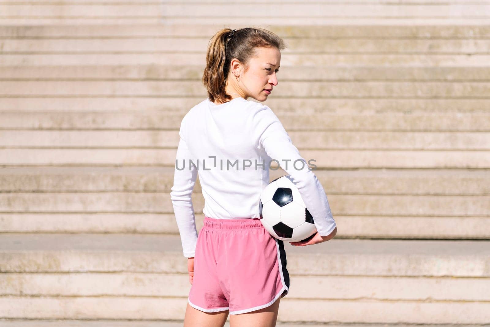 rear view of a sports woman in a concrete soccer court with a ball under her arm, concept of football lifestyle and urban sport in the city, copy space for text