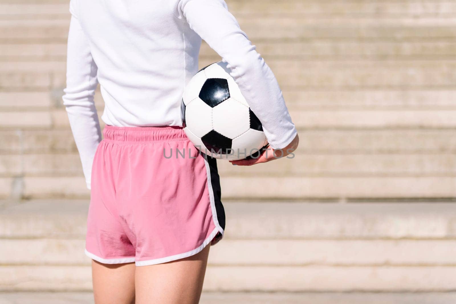 rear view of an unrecognizable sports woman in a concrete soccer court with a ball under her arm, concept of football lifestyle and urban sport in the city, copy space for text