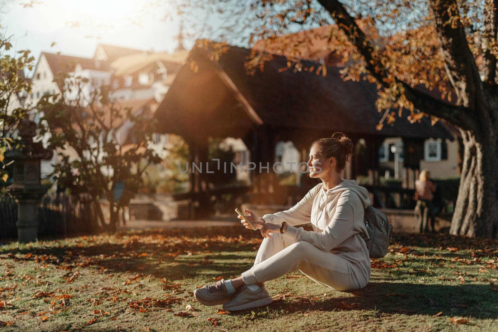 Young Fashionable Teenage Girl With Smartphone In Europian Park In Autumn Sitting At Smiling. Trendy Young Woman In Fall In Park Texting. Retouched, Vibrant Colors. Beautiful Blonde Teenage Girl Wearing Casual Modern Autumn Outfit Sitting In Park In Autumn. Retouched, Vibrant Colors, Brownish Tones. by Andrii_Ko