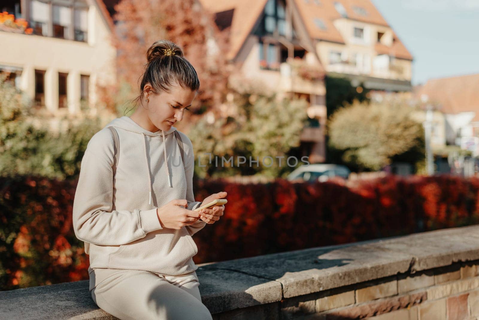 Young Fashionable Teenage Girl With Smartphone In Europian Park In Autumn Sitting At Smiling. Trendy Young Woman In Fall In Park Texting. Retouched, Vibrant Colors. Beautiful Blonde Teenage Girl Wearing Casual Modern Autumn Outfit Sitting In Park In Autumn. Retouched, Vibrant Colors, Brownish Tones. by Andrii_Ko