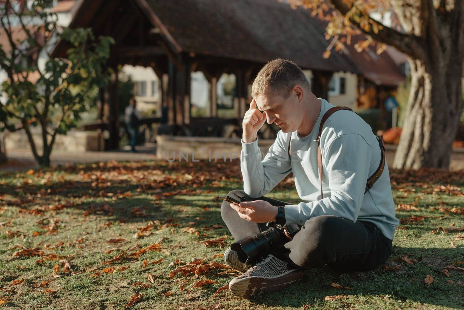 Professional Photographer Taking Picture Of Beautiful Autumn Park. Man Professional Photographer Sit With Camera And With Smartphone In Autumn Park. Retouched, Vibrant Colors, Brownish Tones. by Andrii_Ko