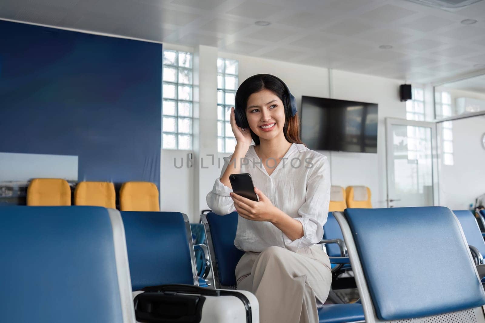 Young Woman Waiting to Board Plane at Airport Terminal, Listening to Music on Headphones and Holding Smartphone by wichayada