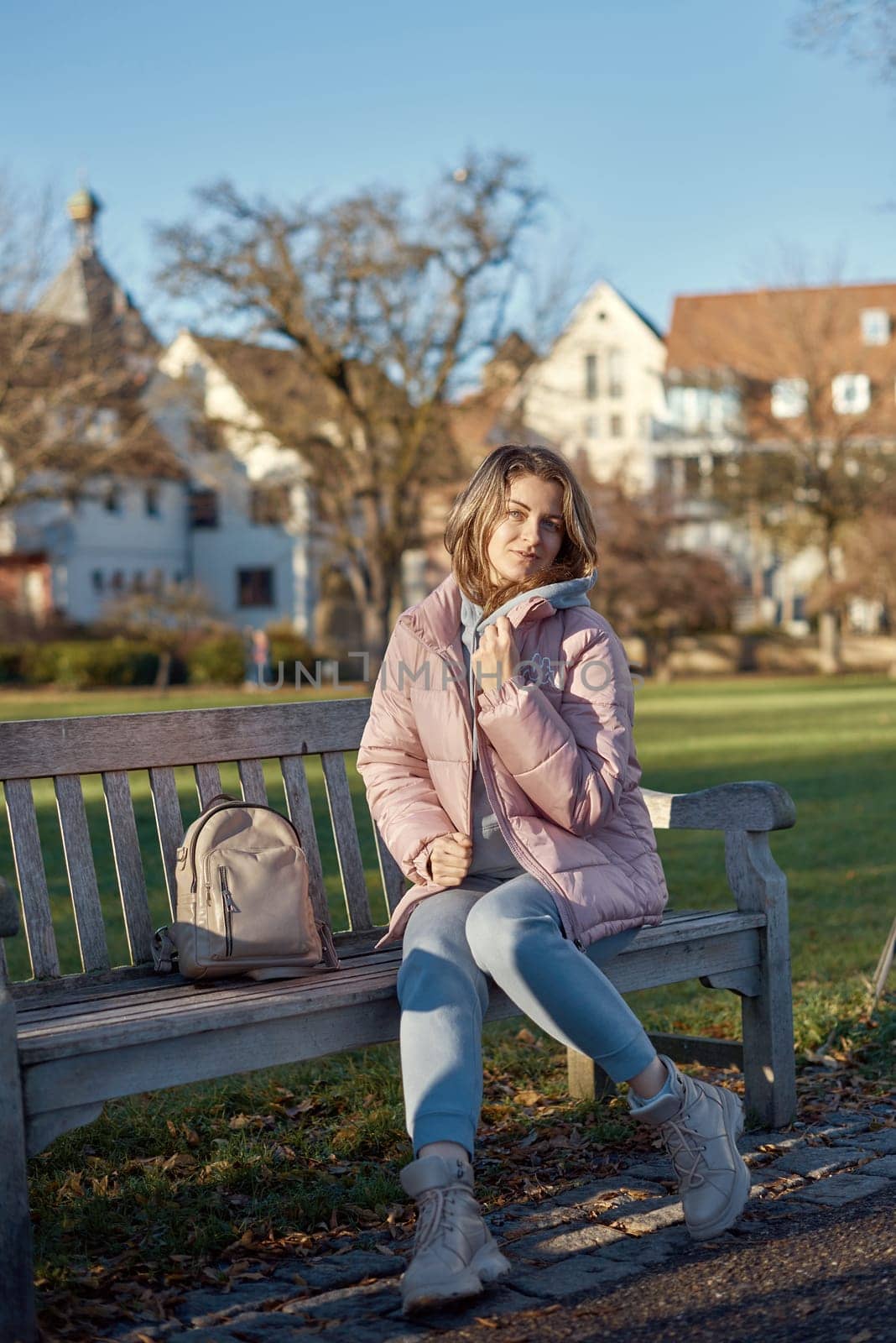 Beautiful Girl in Pink Jacket Relaxing on a Park Bench in the Background of an Old European Town. by Andrii_Ko