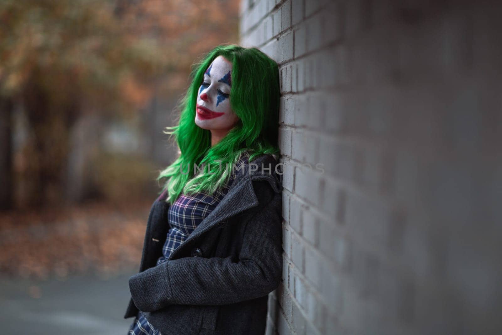 Portrait of a greenhaired girl in chekered dress with joker makeup on a brick wall blurred background.