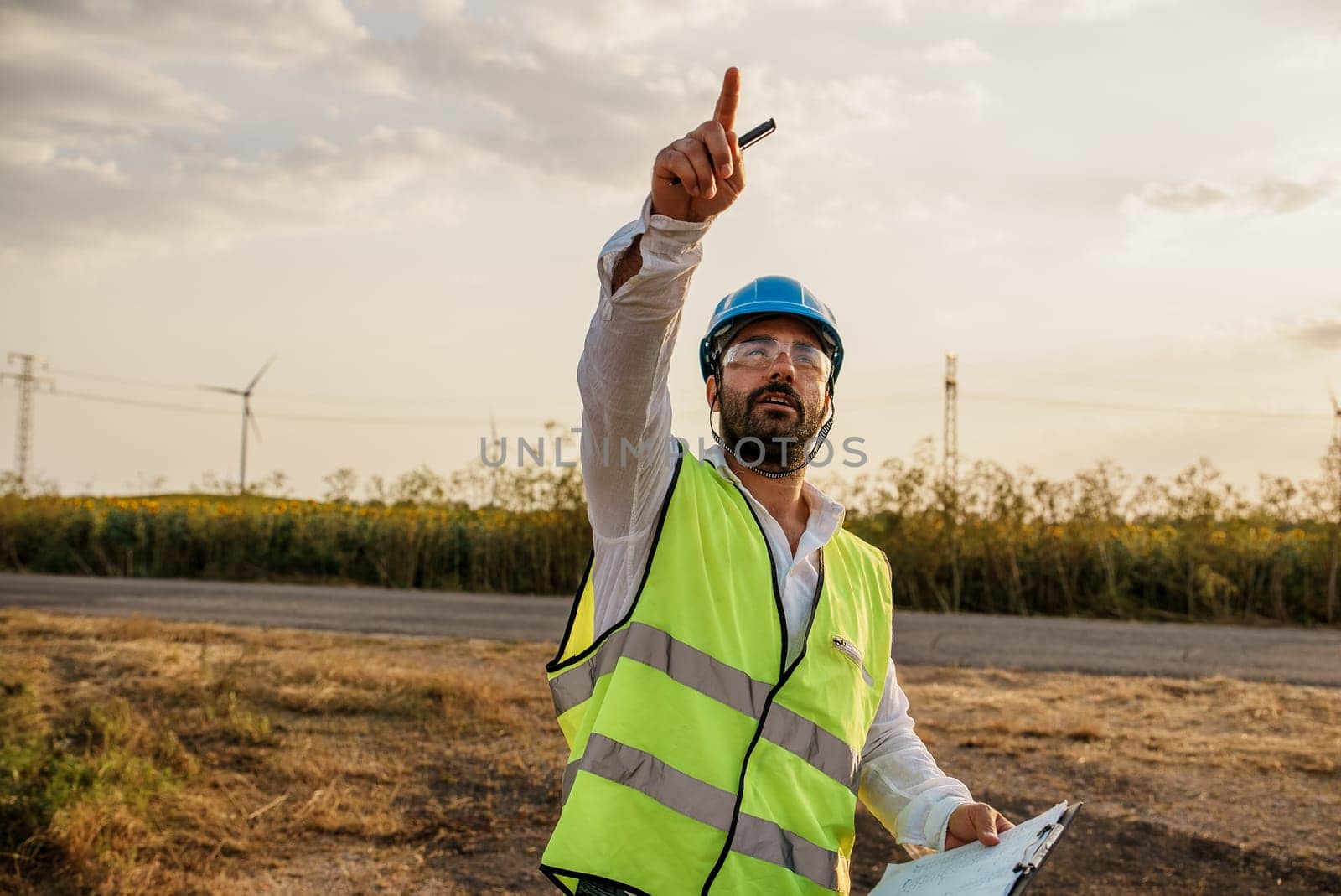 Maintenance engineer points with his hand at a defective turbine in a wind turbine park. Portrait of skilled professional. European green deal.