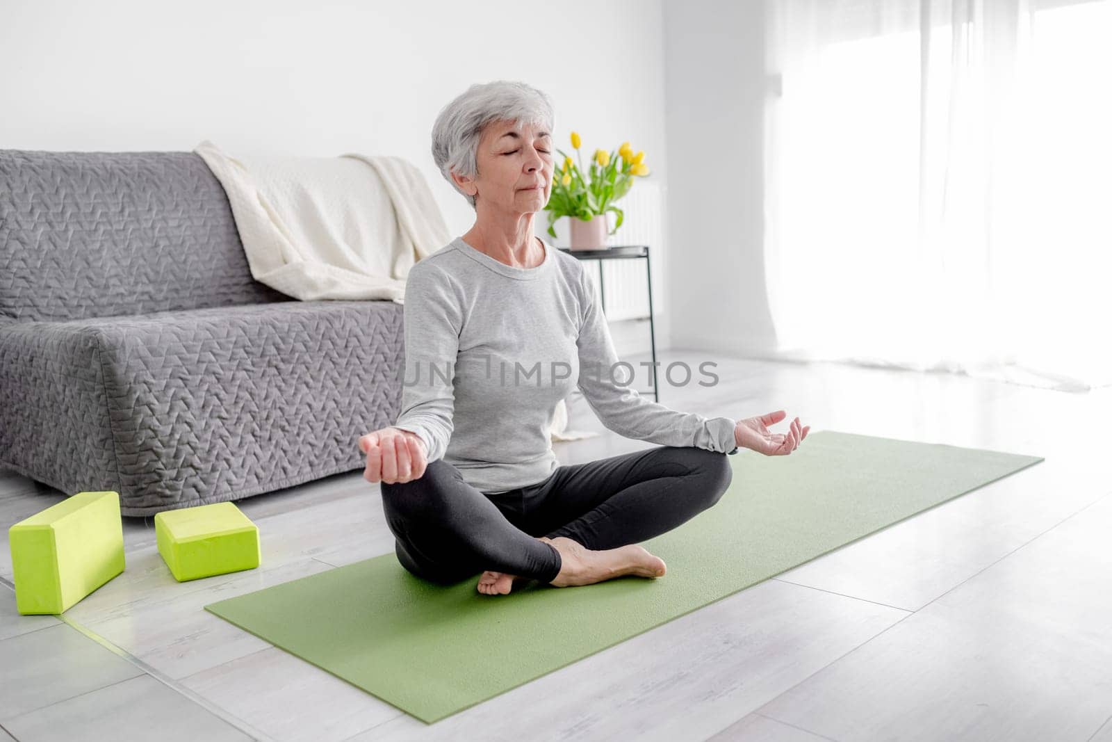 70-Year-Old Woman Practices Yoga In Lotus Pose At Home In A Bright Room