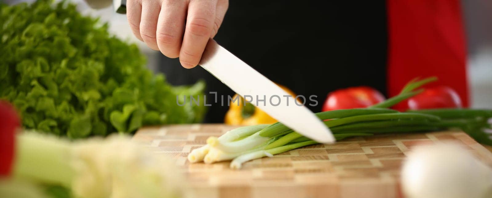 Cook holds knife in hand and cuts on cutting board green onions for salad or fresh vegetable soup with vitamins. Raw food and vegetarian recipe book in modern society popular concept.