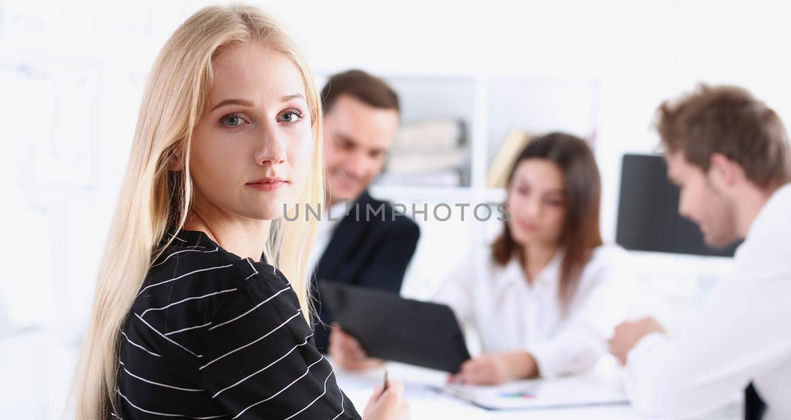 Beautiful smiling cheerful girl at workplace look in camera with colleagues group in background. White collar worker at workspace job offer modern lifestyle client visit profession train concept