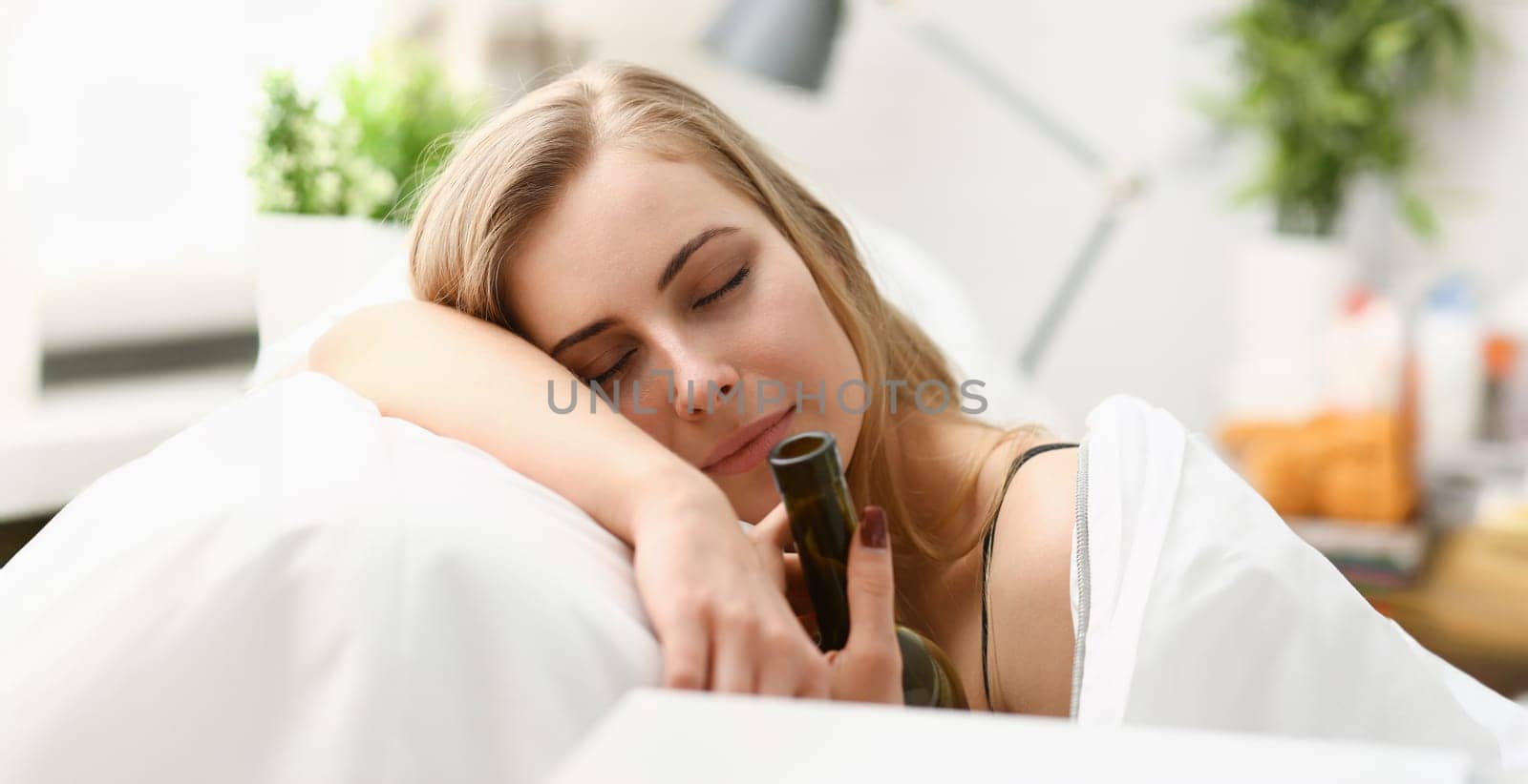 Lonely young woman sleeps early in morning at home embrace with bottle of wine in bedrooms bed with white sheets