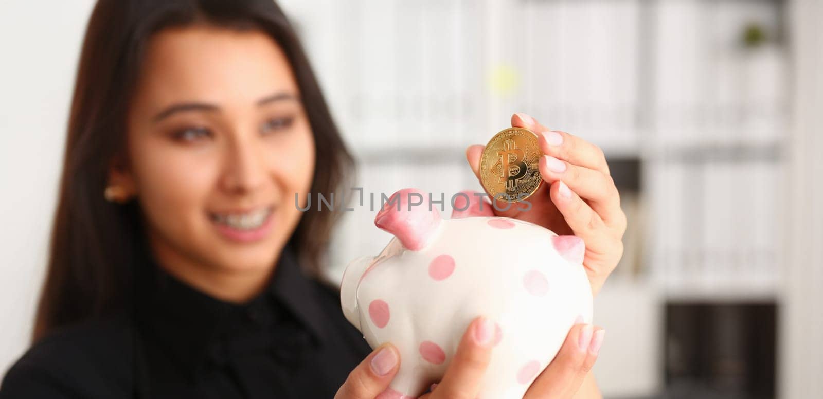A young smiling chinese woman holding bitcoin coin in his hand thrusts it into piggy bank form of pink piggy saving storage theme in crypto currency.