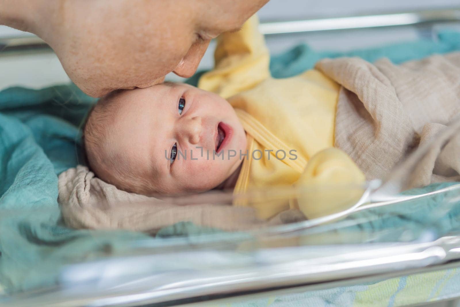 Mom kisses her newborn baby who is resting in a transparent cradle in the hospital. This tender moment emphasizes the bond between mother and child, creating a cozy and safe atmosphere for the infant