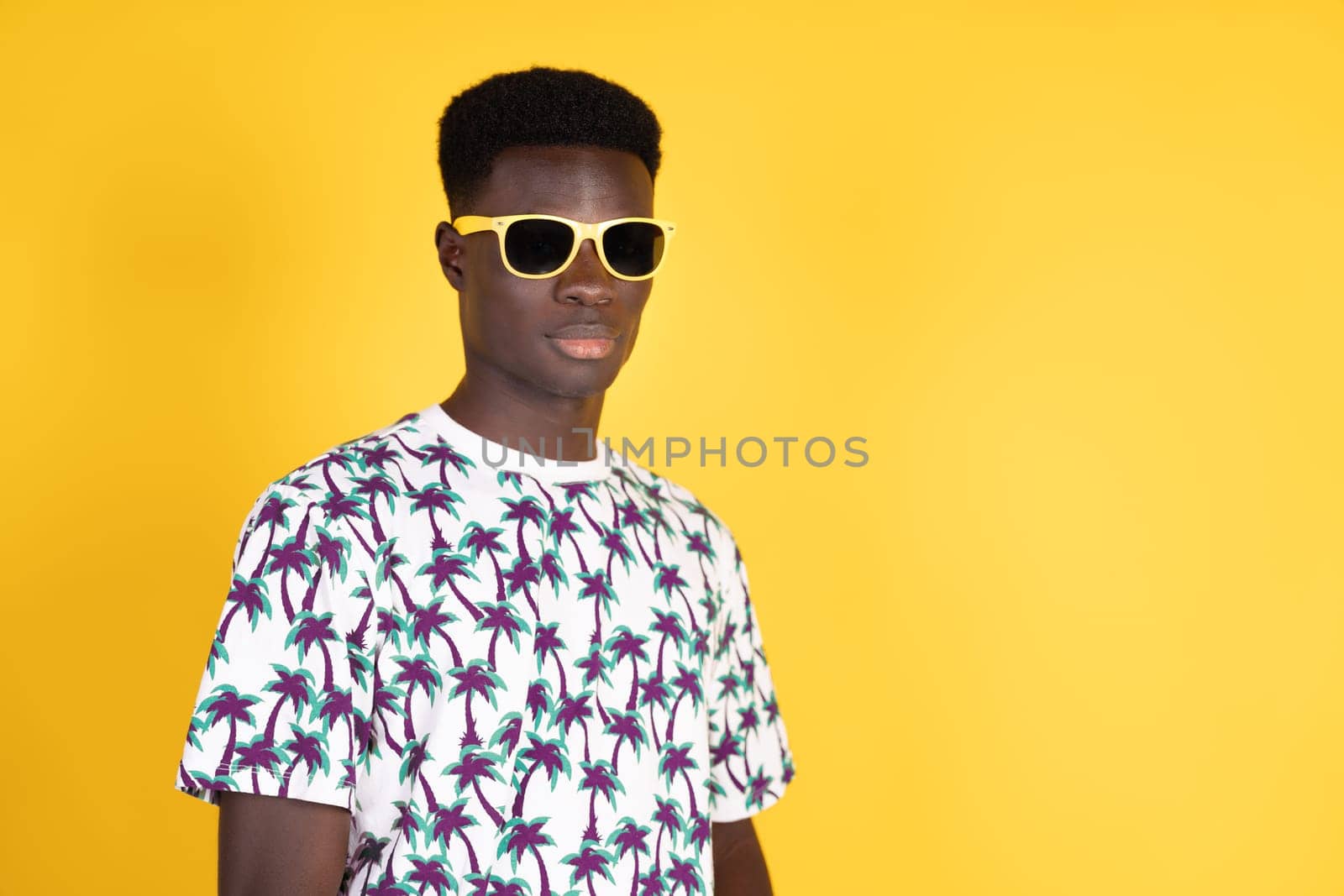 A man wearing a white shirt with palm trees on it stands in front of a yellow background. He is wearing sunglasses and has a serious expression on his face. Concept of relaxation and leisure