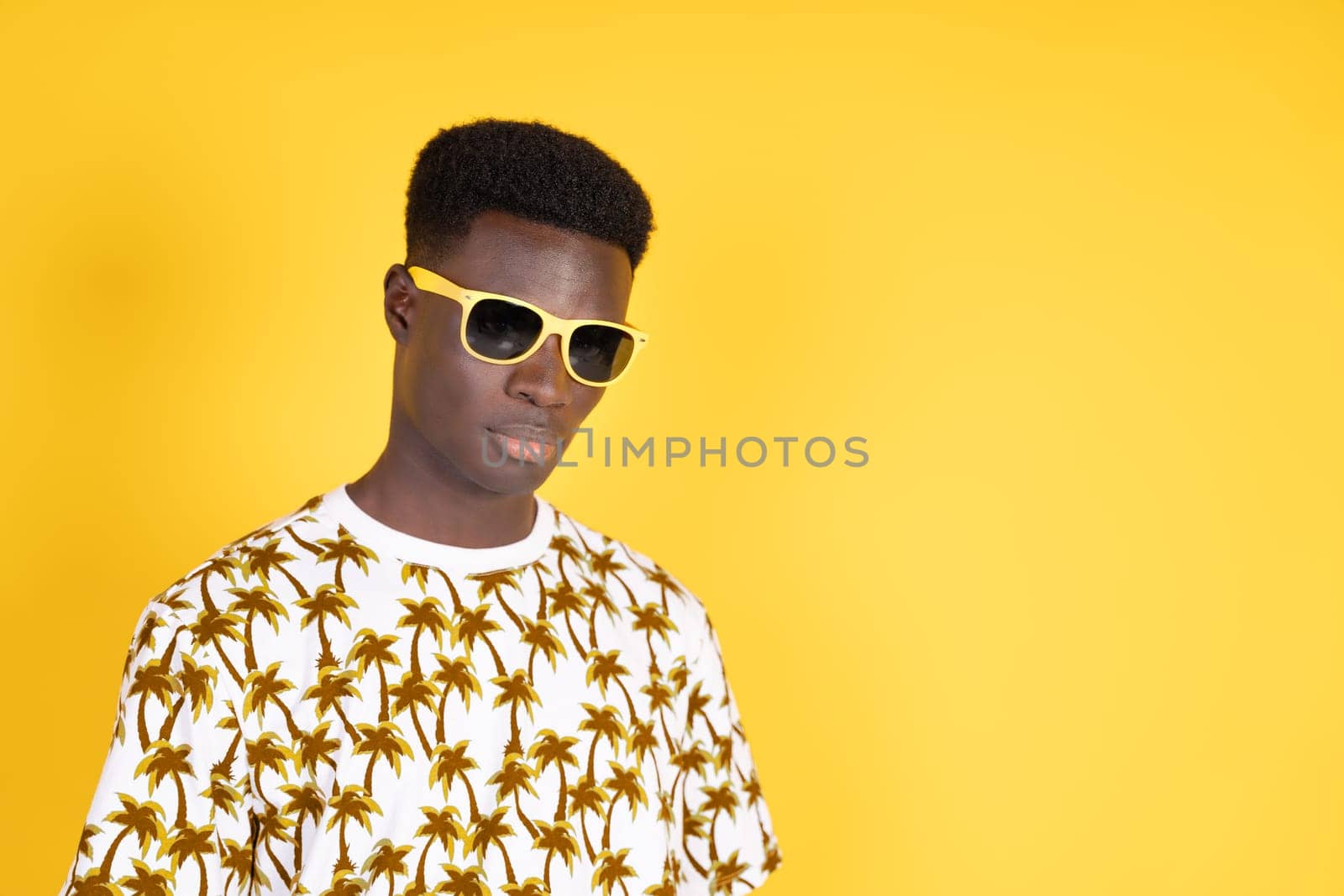 A man wearing sunglasses and a yellow shirt with palm trees on it. He is standing in front of a yellow wall