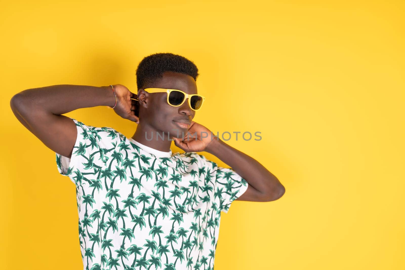 Young black man in yellow sunglasses and summer t-shirt with palm trees on a yellow background with copy space. by Ceballos