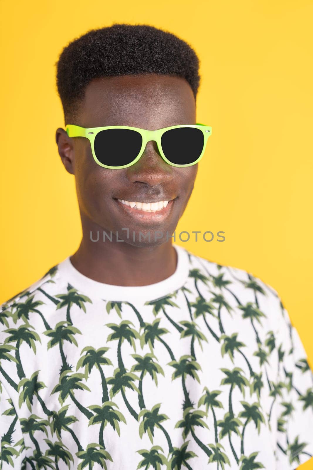 A man with a green pair of sunglasses on his face. He is smiling and wearing a shirt with palm trees on it