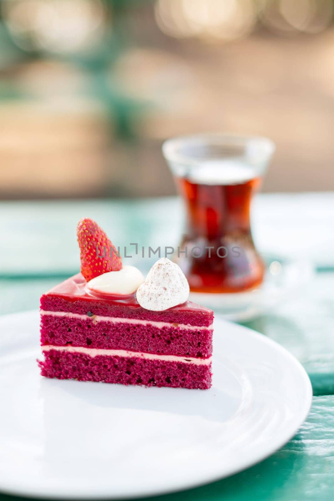 piece of red velvet cake decorated with fresh berries in a white plate on the table, a cup of Turkish tea in a traditional Armudu glass, time for a sweet dessert during the summer holidays